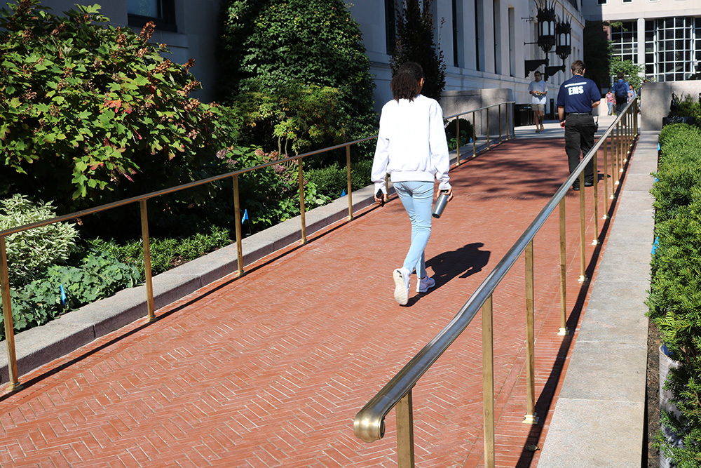 Student wearing a white sweatshirt and blue jeans walks up an access ramp. The ramp is made of brick pavers and has gold railings on either side.