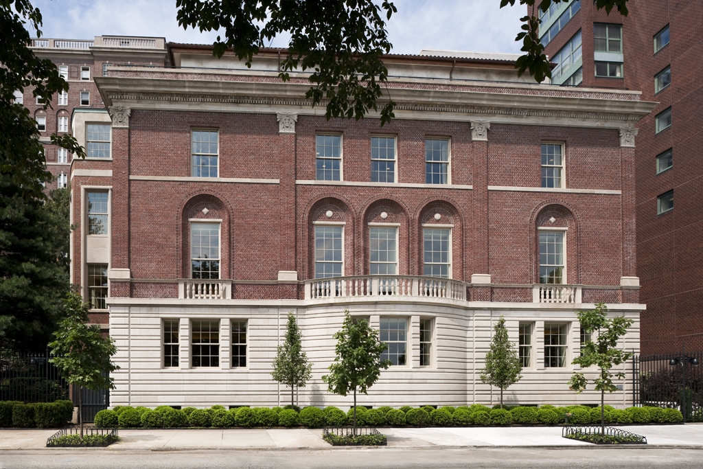 A 6-story red brick and limestone building. The second floor has floor-to-ceiling windows and a balcony. In front of the building is a shrub-lined sidewalk.