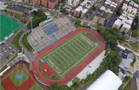 An aerial view of a green, Columbia-branded turf football field surrounded by an orange rubber track. The fieldis part of a stadium complex. Around the football field are partial views of a turf soccer field and a baseball field.