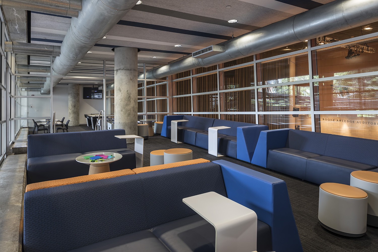 Lounge area with navy blue couches and small round stools. The lunge is divided by a partial glass wall with study tables in the other section.