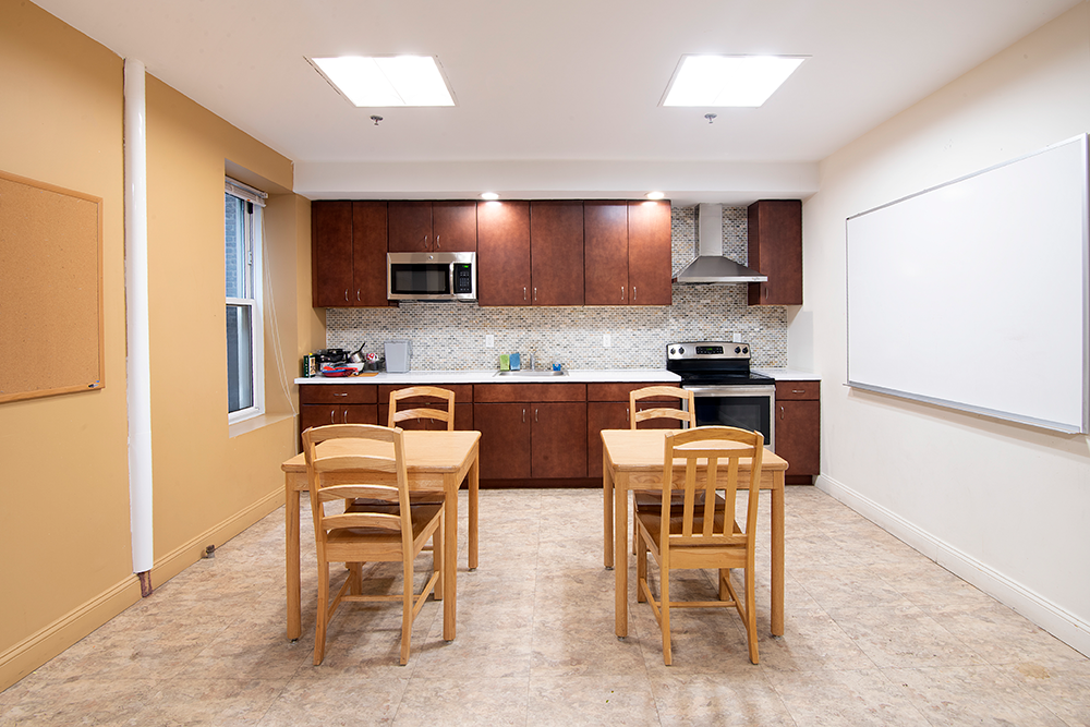 A joint kitchen and dining space. Two wooden tables are situated in front of a kitchen with white countertops, dark, wooden cabinets, and a stainless steel range and microwave.