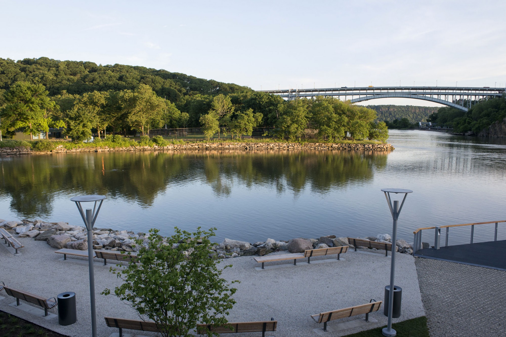 Gravel walkway with wooden benches and grey street lamps. The benches look over a river with a bridge in the background.