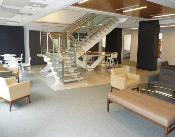 Open collaboration space with white high-top tables and stools and neutral-colored lounge chairs and couches. A staircase is in the center of the space.