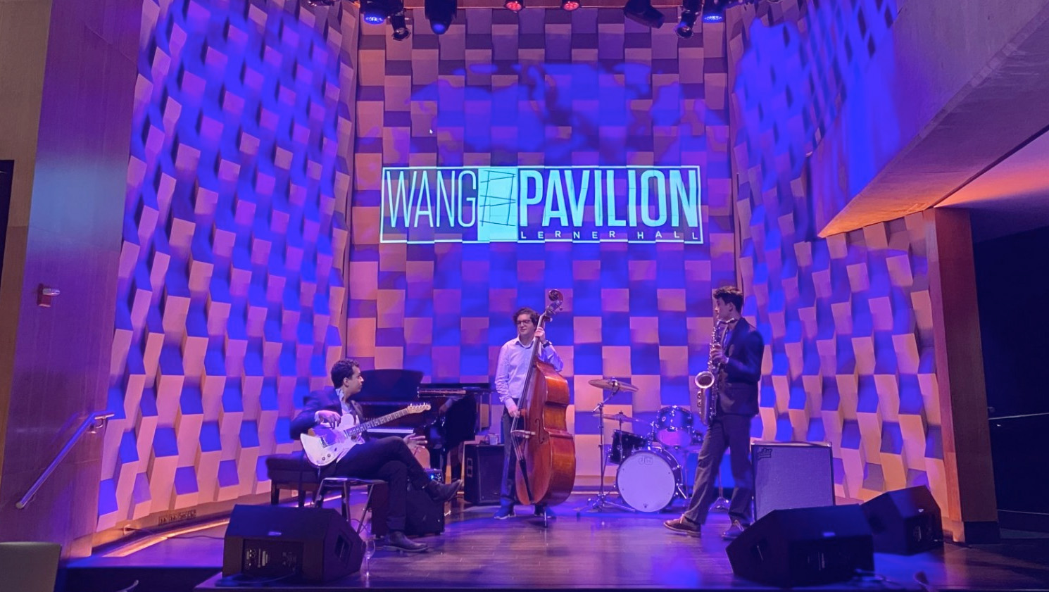 A jazz trio jams out on the stage of the Wang Pavilion, which has purple and pink lights illuminating the wall behind them.