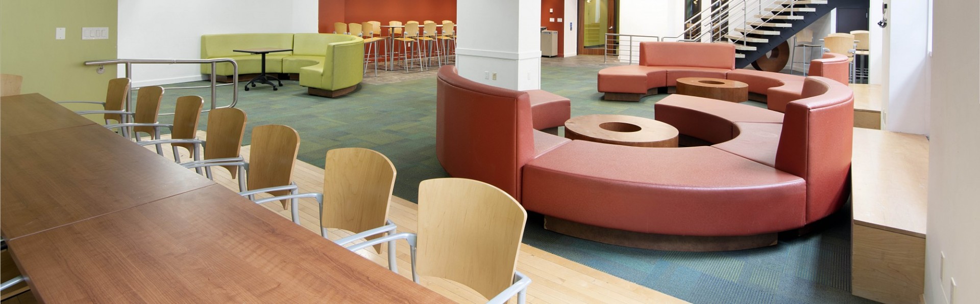 An empty residential building lounge with circular red and green couches and long wooden tables with wooden chairs.