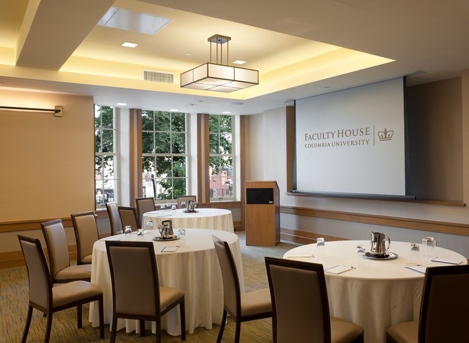 Round tables dressed with white linens facing a projector screen that says, "Faculty House." Each table is surrounded by square-backed, grey chairs. 