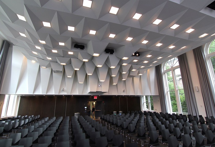(Photo: Architect – LTL Architects; Michael Schissel, photographer) Large, white-walled lecture hall with rows of black plastic chairs and floor-to-ceiling windows. The custom ceiling is designed to accommodate acoustics. 