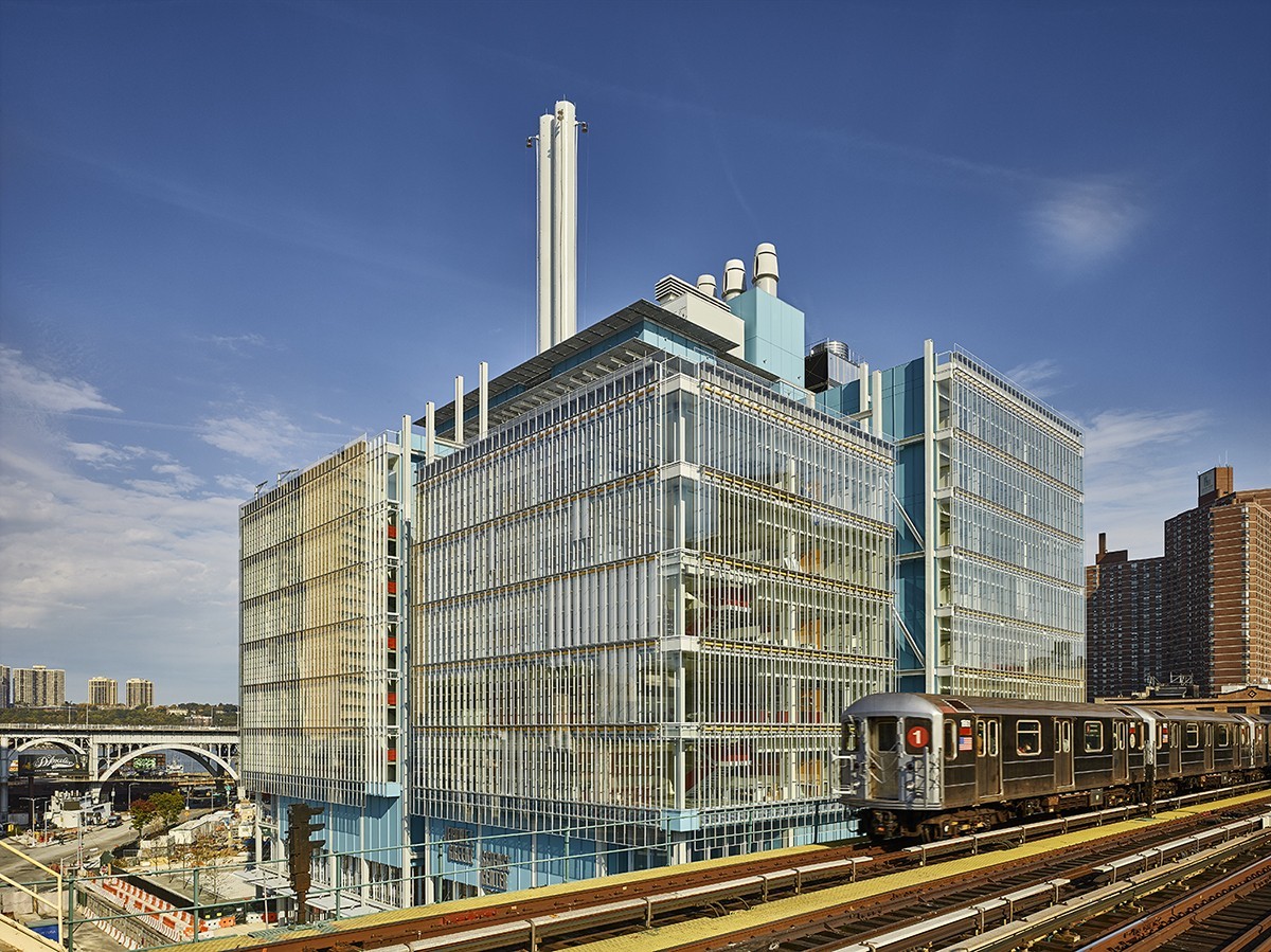 Exterior view of the Jerome L. Greene Science Center, looking northwest. The building is a steel and glass structure. A subway train moves along tracks in the foreground of the photo.