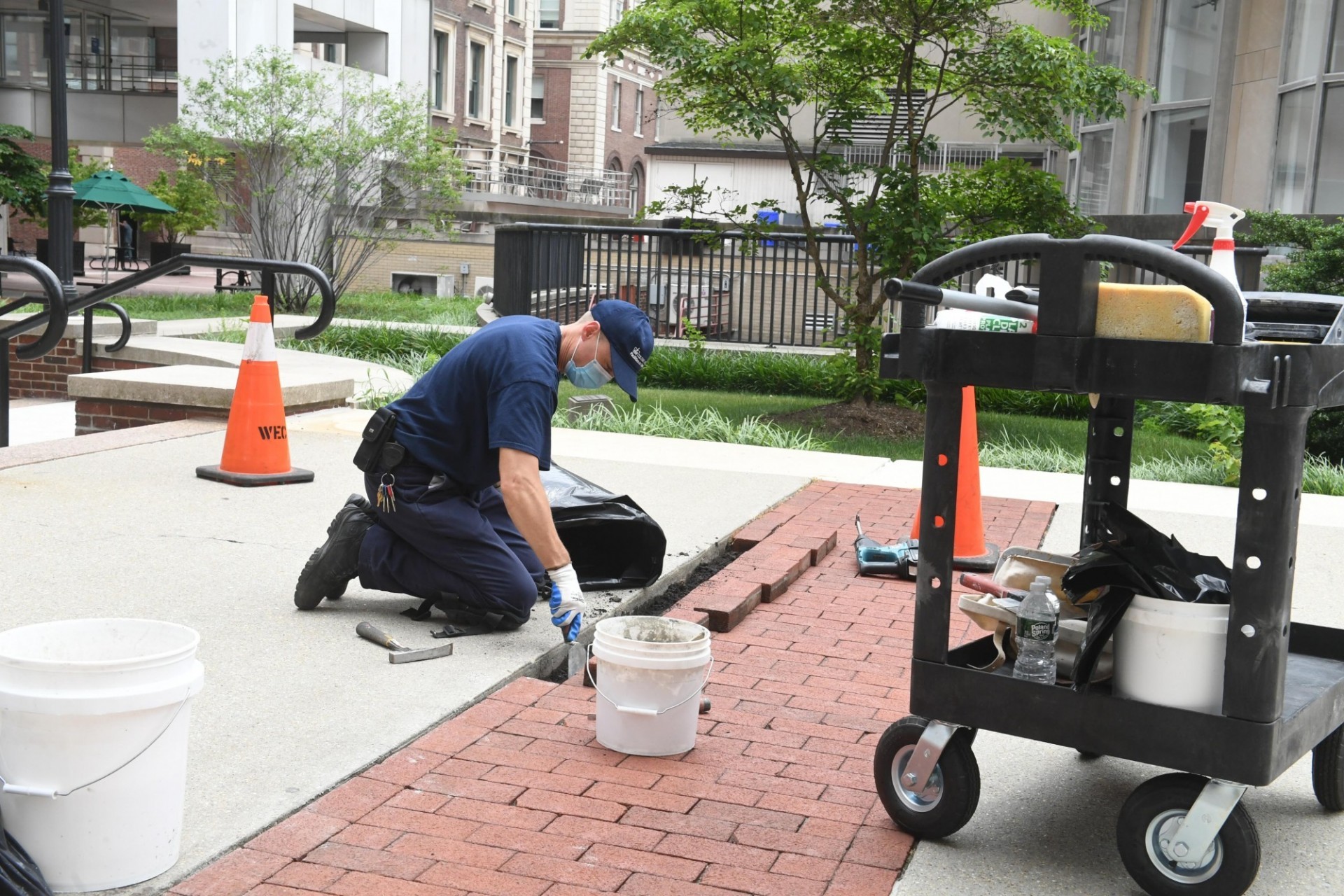 A member of the Facilities and Operations team wearing a dark blue t-shirt and baseball cap is replacing brick pavers in a pathway.