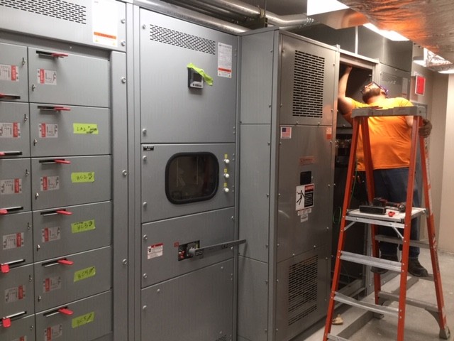 Facilities worker in orange shirt and blue jeans works on a large scale circuit breaker system.
