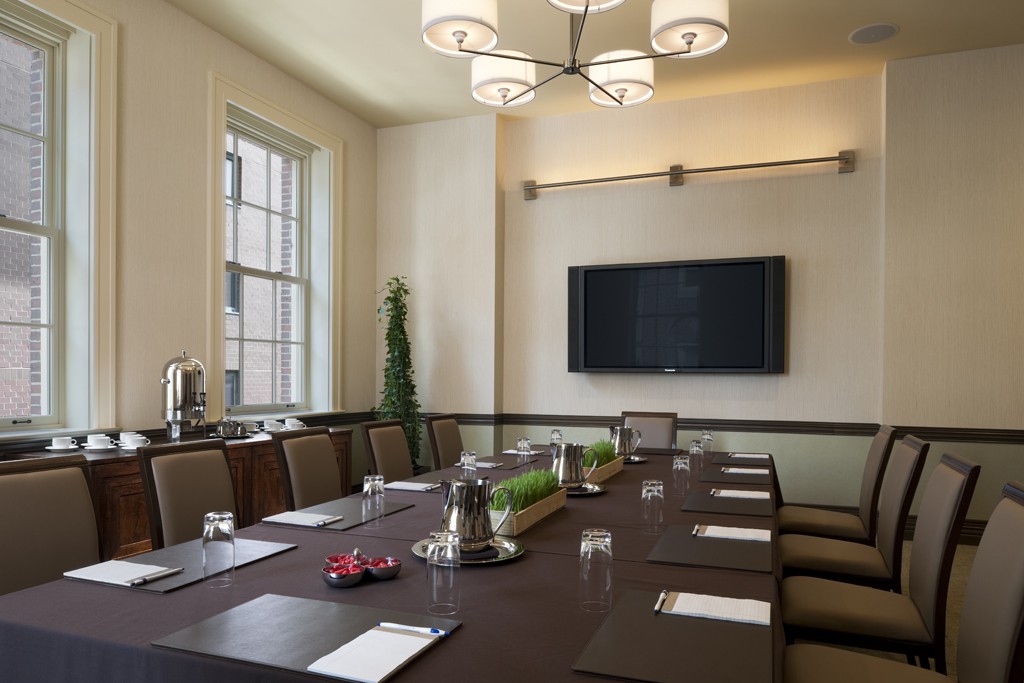 A meeting room with a long, mahogany conference table and brown chairs. At the end of the room, a TV screen is mounted on the wall.