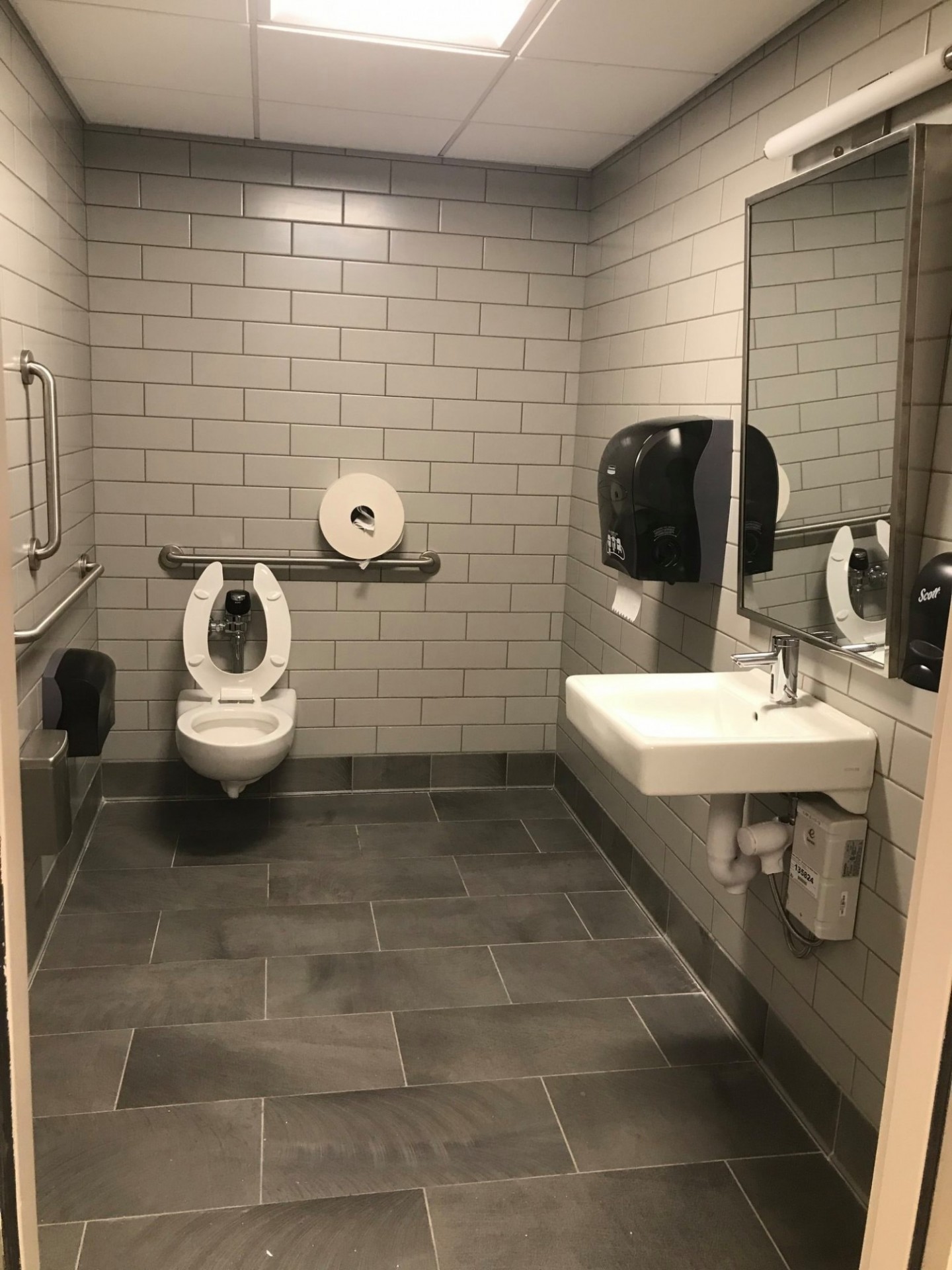 Accessible, single-person restroom with grab bars, white tiled walls, sink, mirror, and gray floor tiles