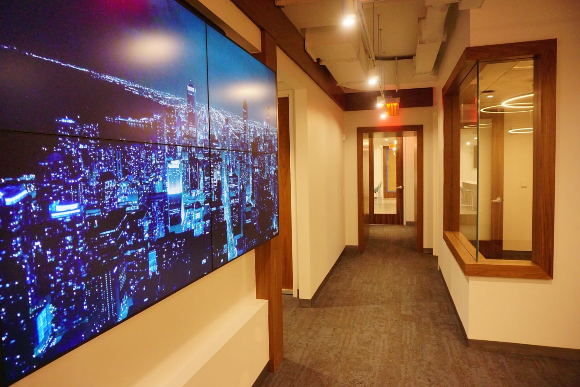 A brown-carpeted hallway with a glass door at the end of it. On the left wall, there is a video screen displaying a city skyline at night.