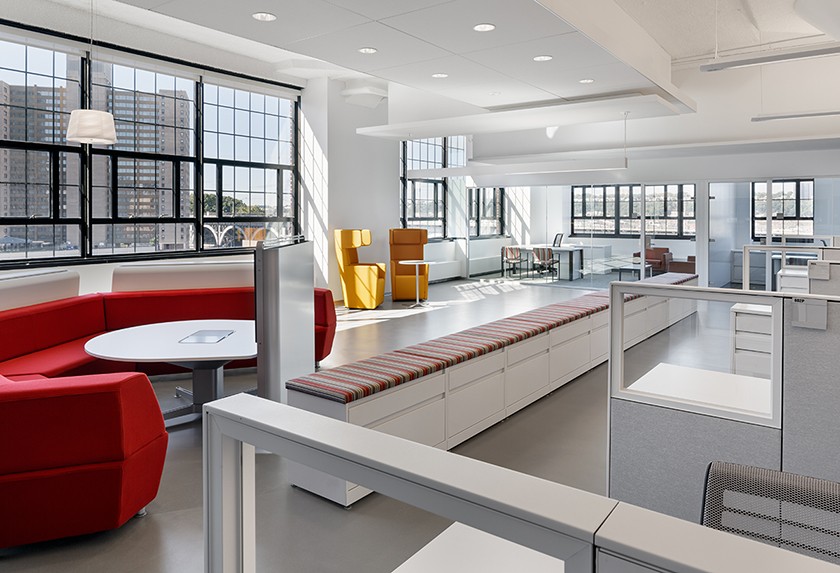 (c) Colin Miller. Renovated, open floor layout with large windows along perimeter, red couch and seating area around a monitor on the left, a long brown bench running the width of the floor, and open cubicles with glass dividers. 