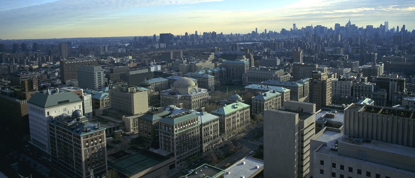 A south-facing aerial view of campus showing University buildings and the Manhattan skyline against a blue sky.