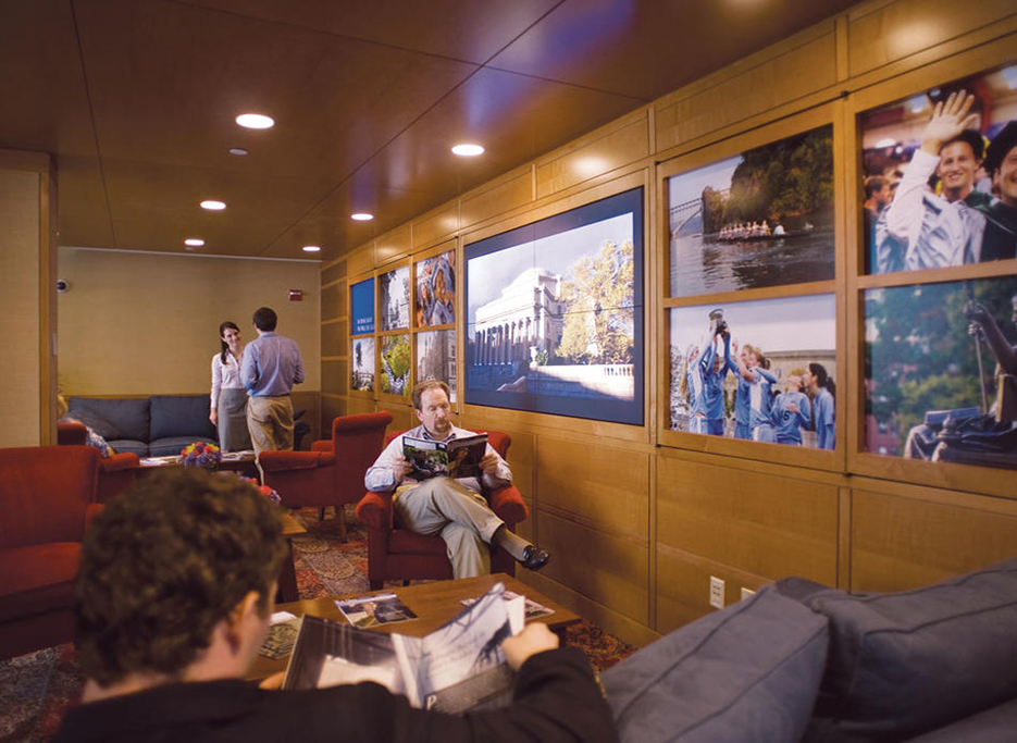 A wood-paneled lounge in use by two men reading magazines and a man and woman in the background having a conversation.