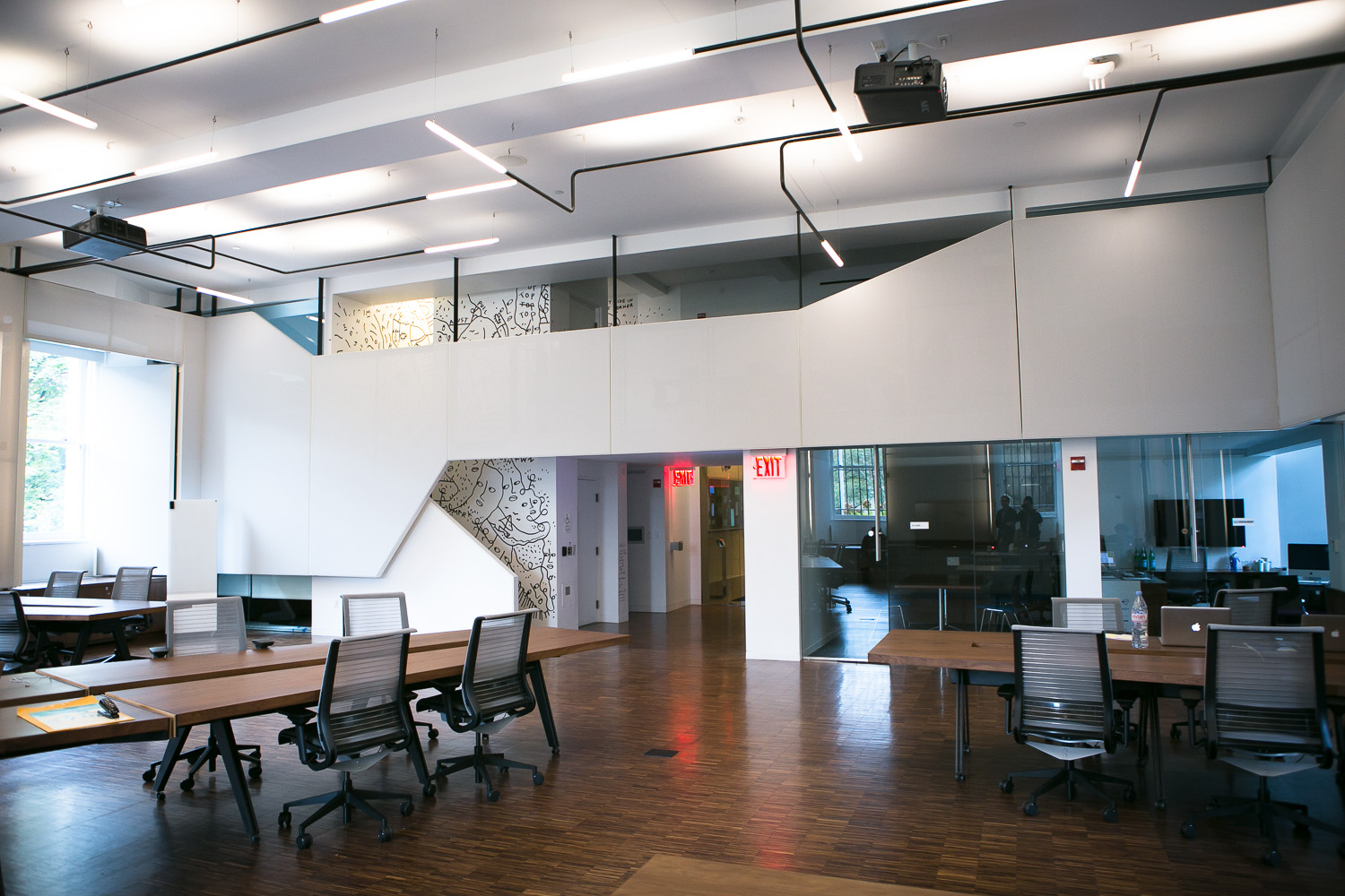 Large, open room with wooden floors, large windows, and white walls. There are multiple long, wooden tables with chairs. 