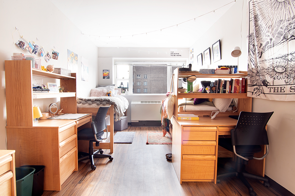 A sunlit residence hall room with white walls. on the left, a wooden desk is pushed up against the wall, parallel to a single bed. On the right, a wooden desk is facing a single bed.