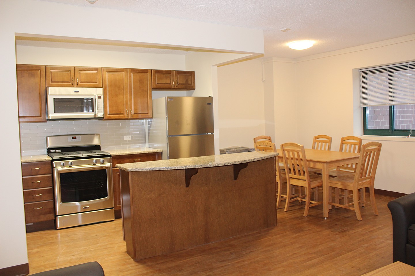 Open-plan kitchen with speckled gray marble countertops, stainless steel fridge, electric stainless steel over, white microwave, and brown wooden cabinets. Next to the kitchen area is a wooden dining table. 