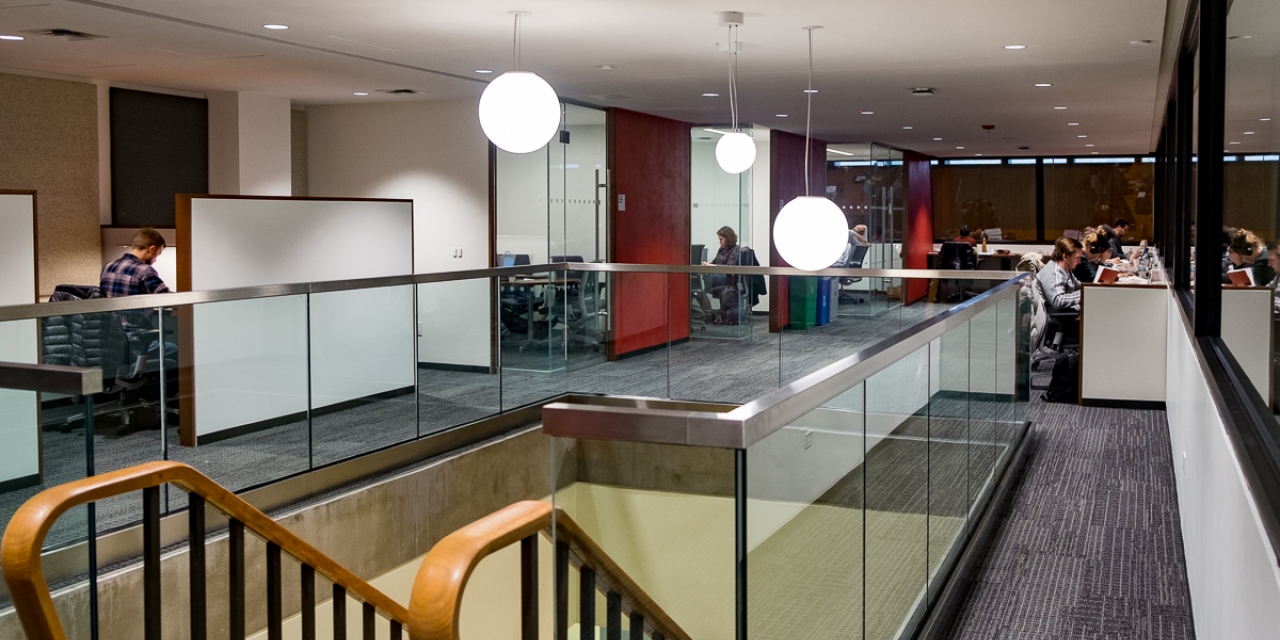 Grey-carpeted workspace area with individual cubicles, private meeting rooms, and open work tables