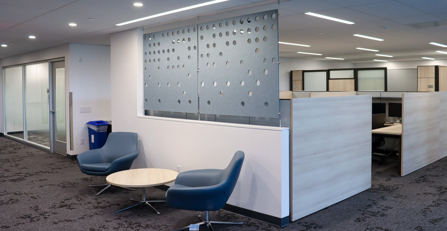 A view of the renovated suite on the fifth floor of the Interchurch Building, with a gray accent wall with holes in it, blue chairs and a coffee table, and wood accent cubicles. 