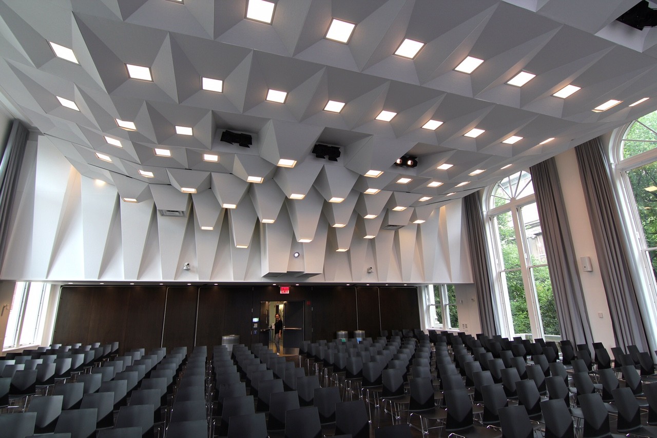 (Photo: Architect – LTL Architects; Michael Schissel, photographer) Large, white-walled lecture hall with rows of black plastic chairs and floor-to-ceiling windows. The custom ceiling is designed to accommodate acoustics. 