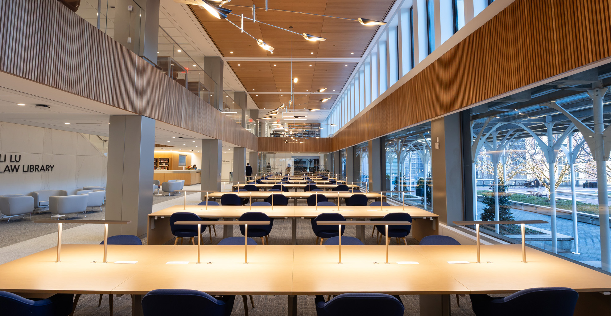 The Li Lu Law Library, a newly renovated library in Jerome Greene Hall, with long tables and blue chairs, and wood panel floor panels and ceiling.