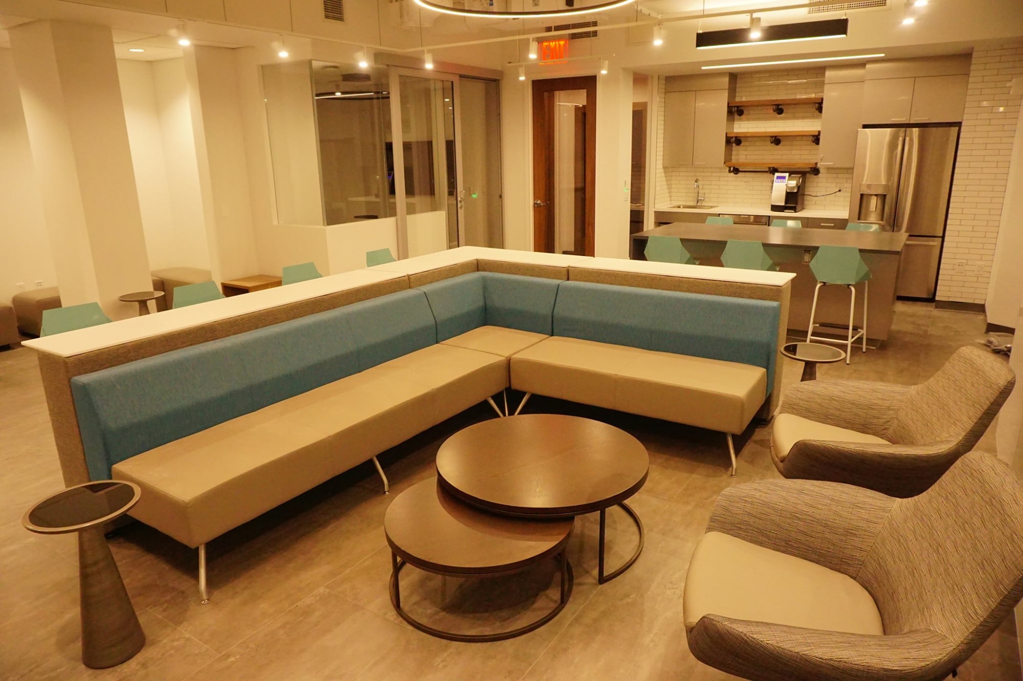 In the foreground, there are two brown arm chairs across from a blue and brown couch. In the background, there is a countertop and kitchen area with grey cabinets and a stainless steel fridge.