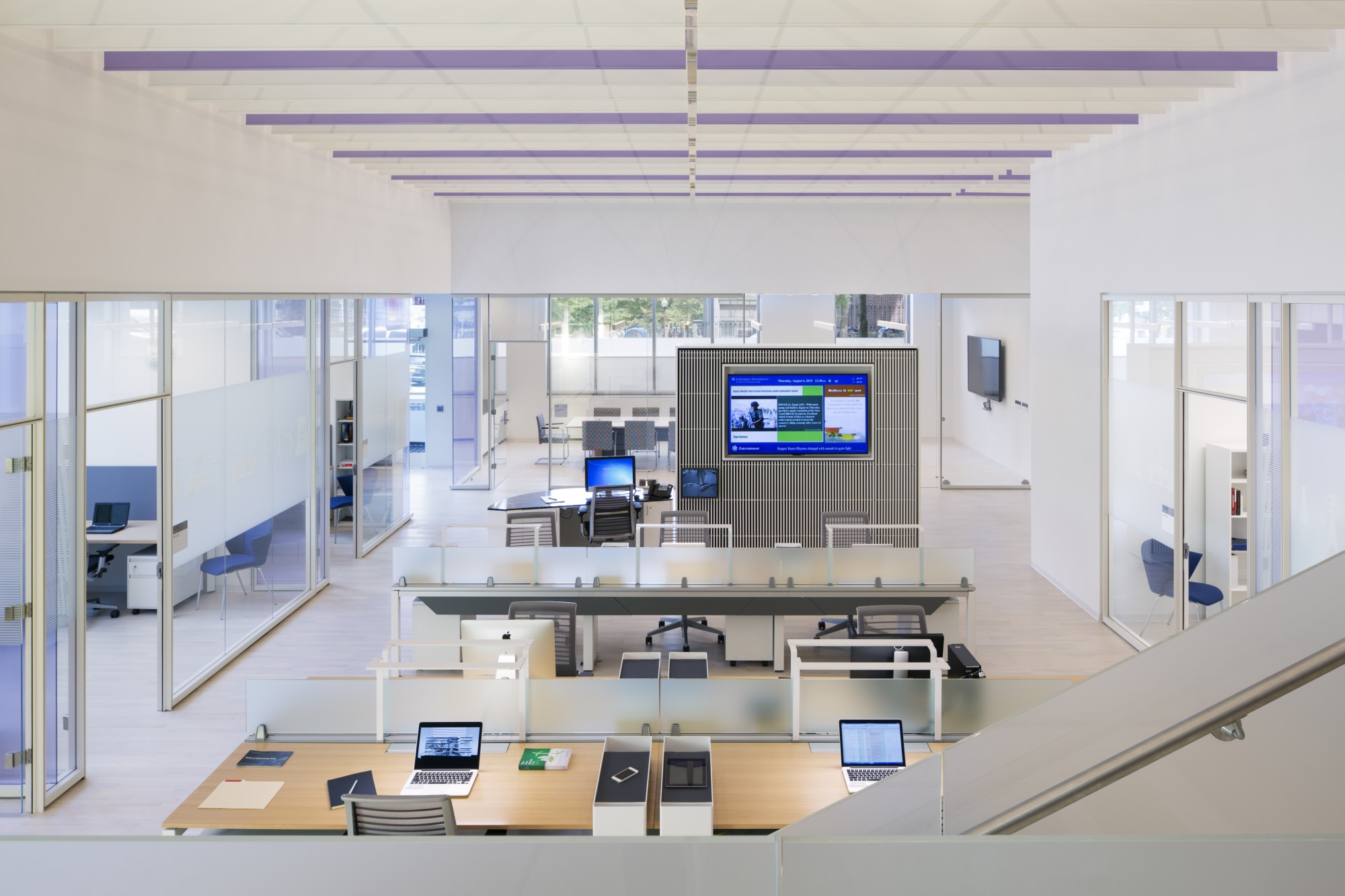 Open work stations with computers and a large screen at the front of the room, with private offices bordering the perimeter