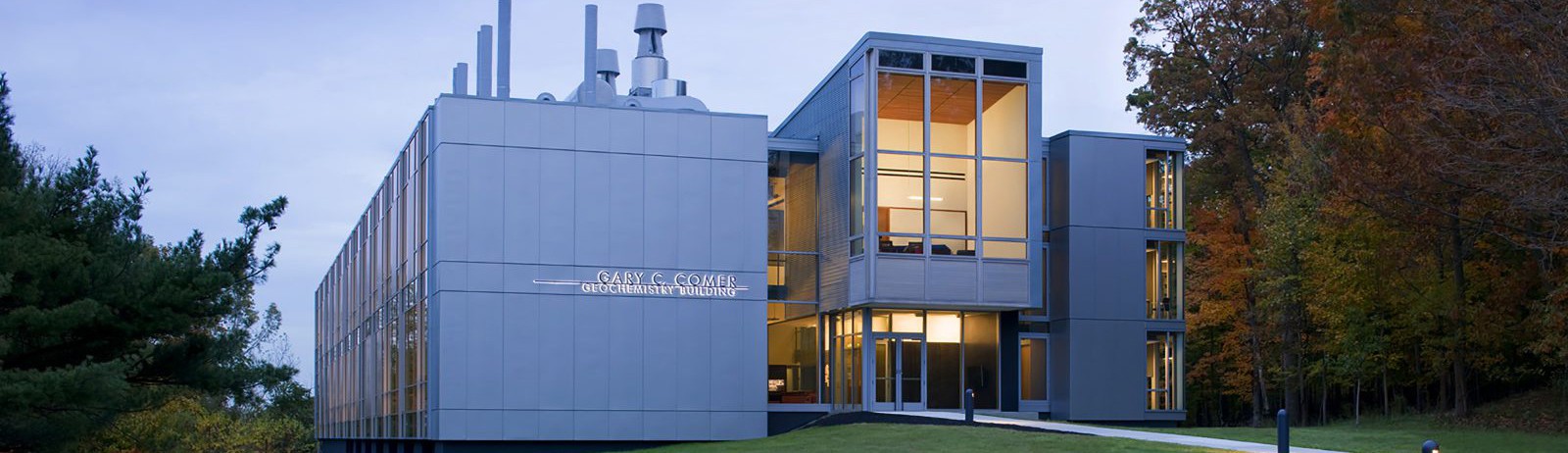 Concrete and glass structure set against sky at dusk. Trees obscure the right sided of the building facade.