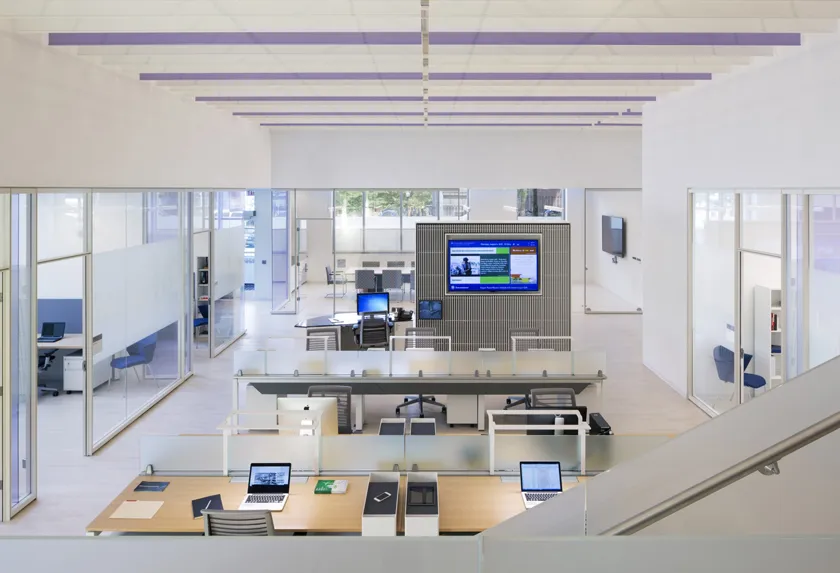 Open work stations with computers and a large screen at the front of the room, with private offices bordering the perimeter