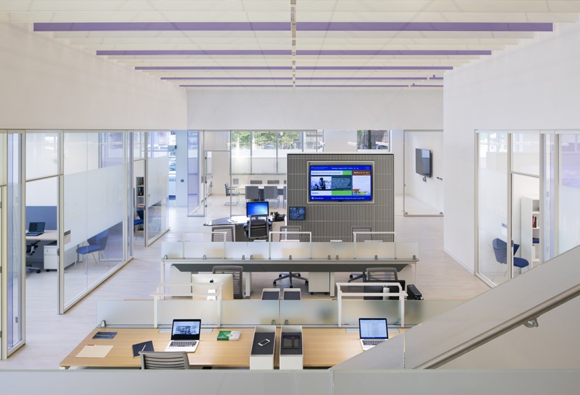 Open work stations with computers and a large screen at the front of the room, with private offices bordering the perimeter