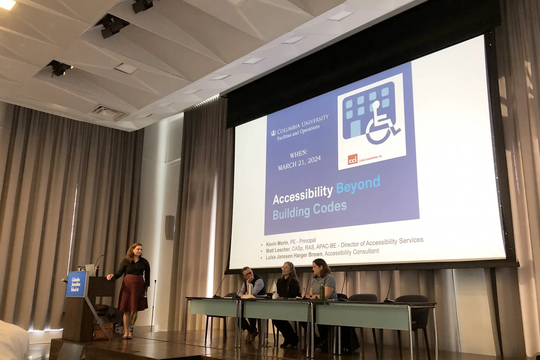 A panel of people sit in front of a projection screen that says, "Accessibility Beyond Building Codes" on the title slide.