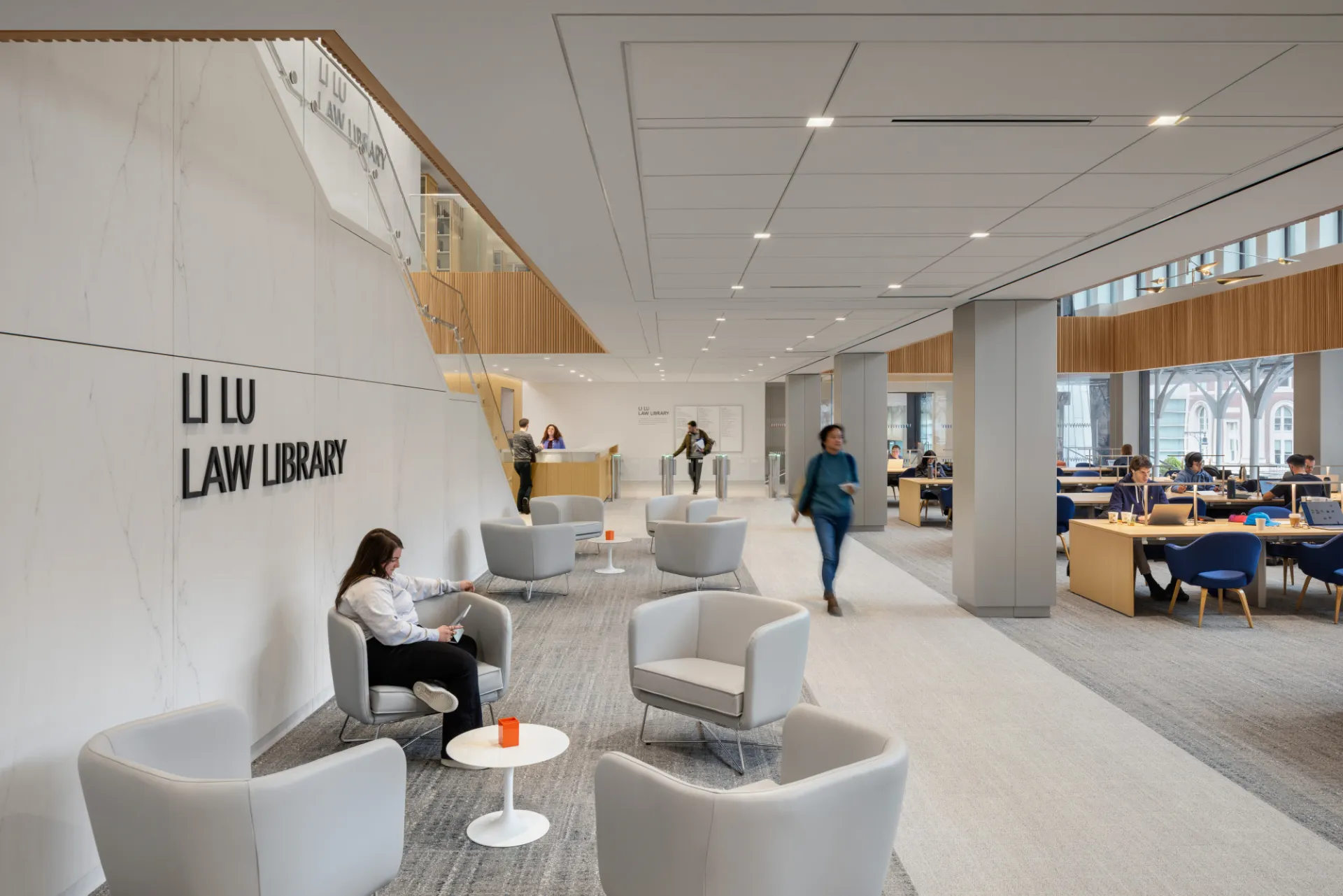 The third floor of the Li Lu Law Library at Jerome Greene Hall, featuring a large reading room on the right with people sitting at long tables, gray chairs and coffee tables on the left wall, and a staircase leading to the upper floor.