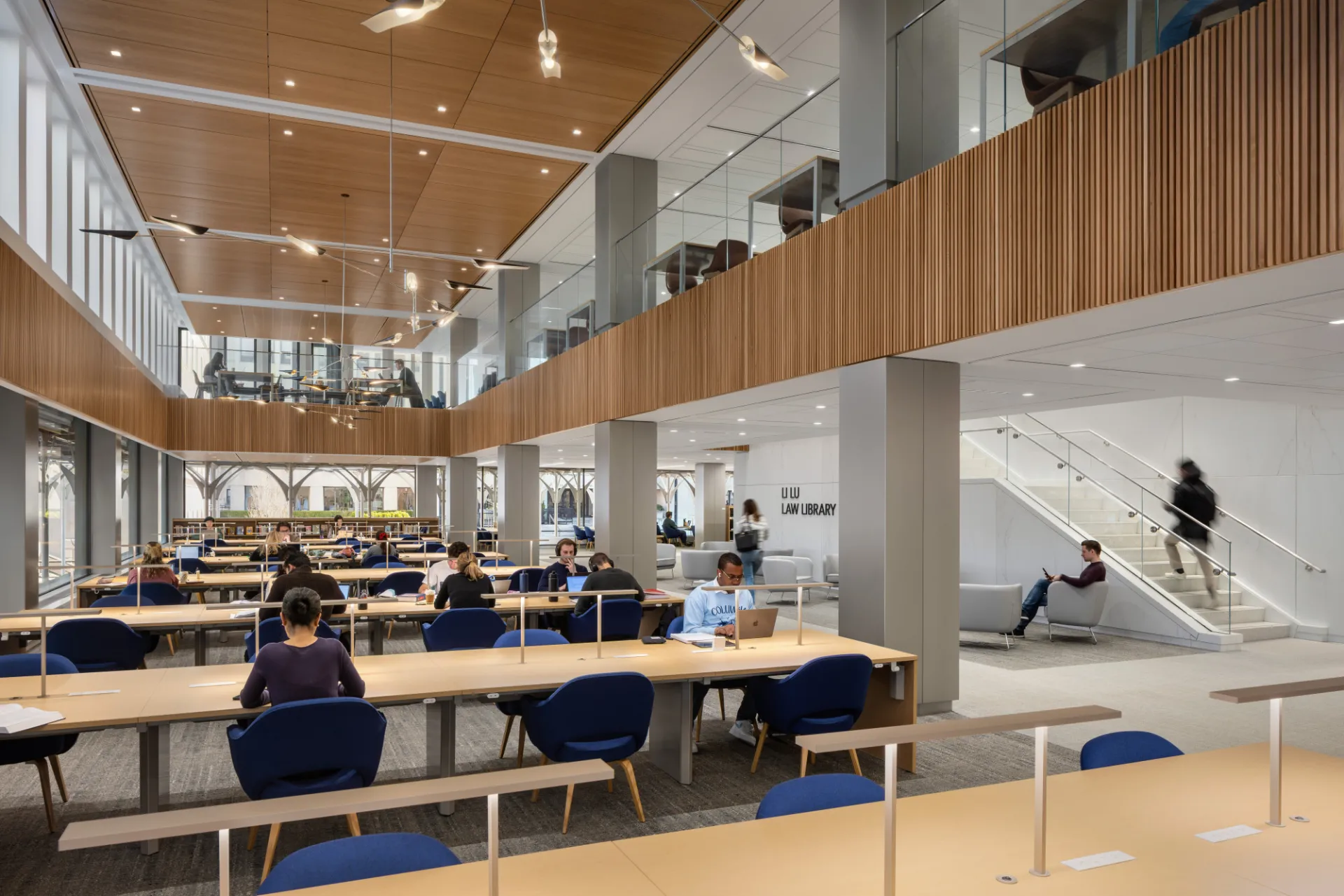 A view of the two-story reading room in the Li Lu Law Library, which has wood accent walls and ceiling, gray columns, long tables, and blue chairs.  