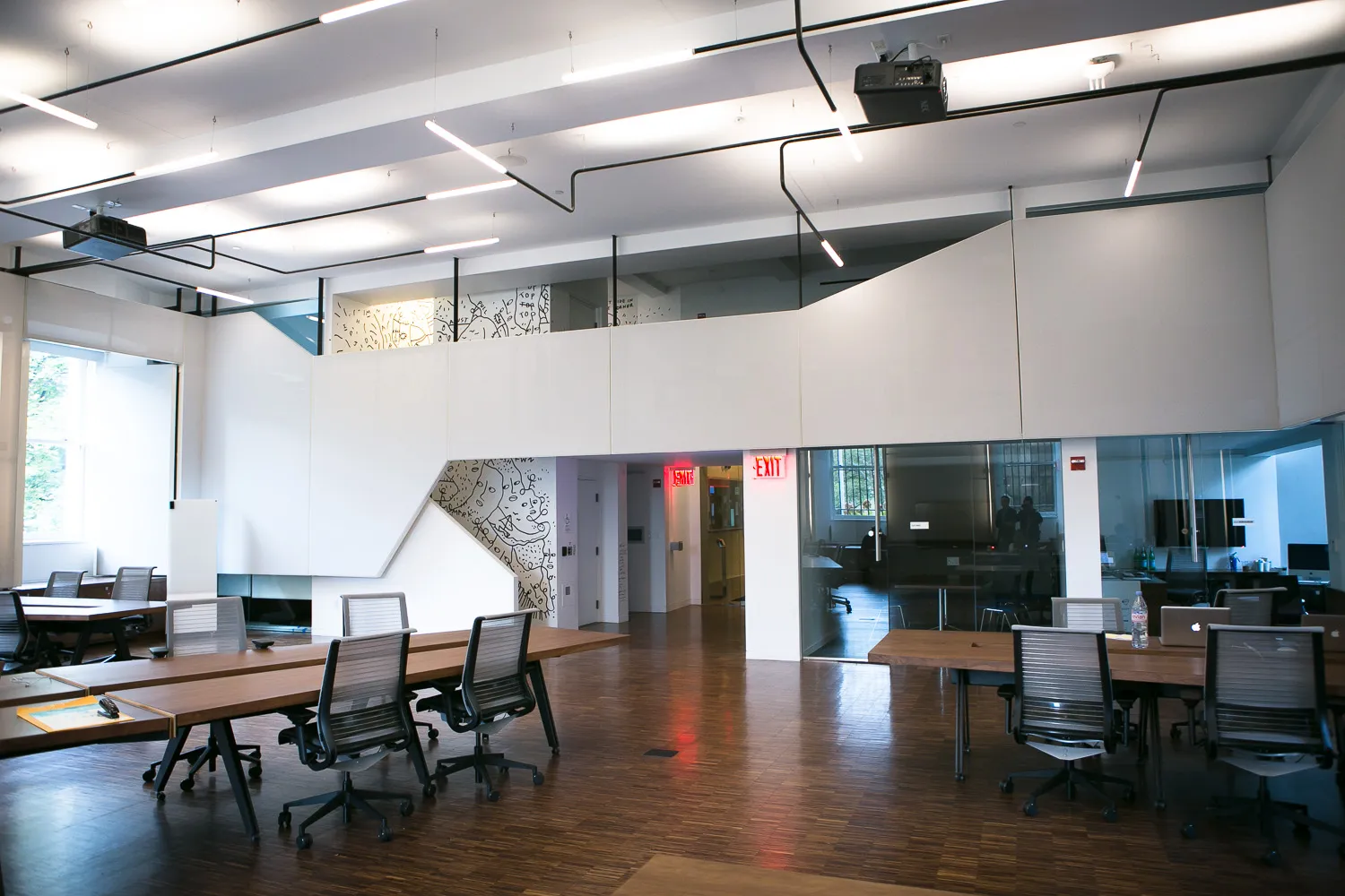 Large, open room with wooden floors, large windows, and white walls. There are multiple long, wooden tables with chairs. 