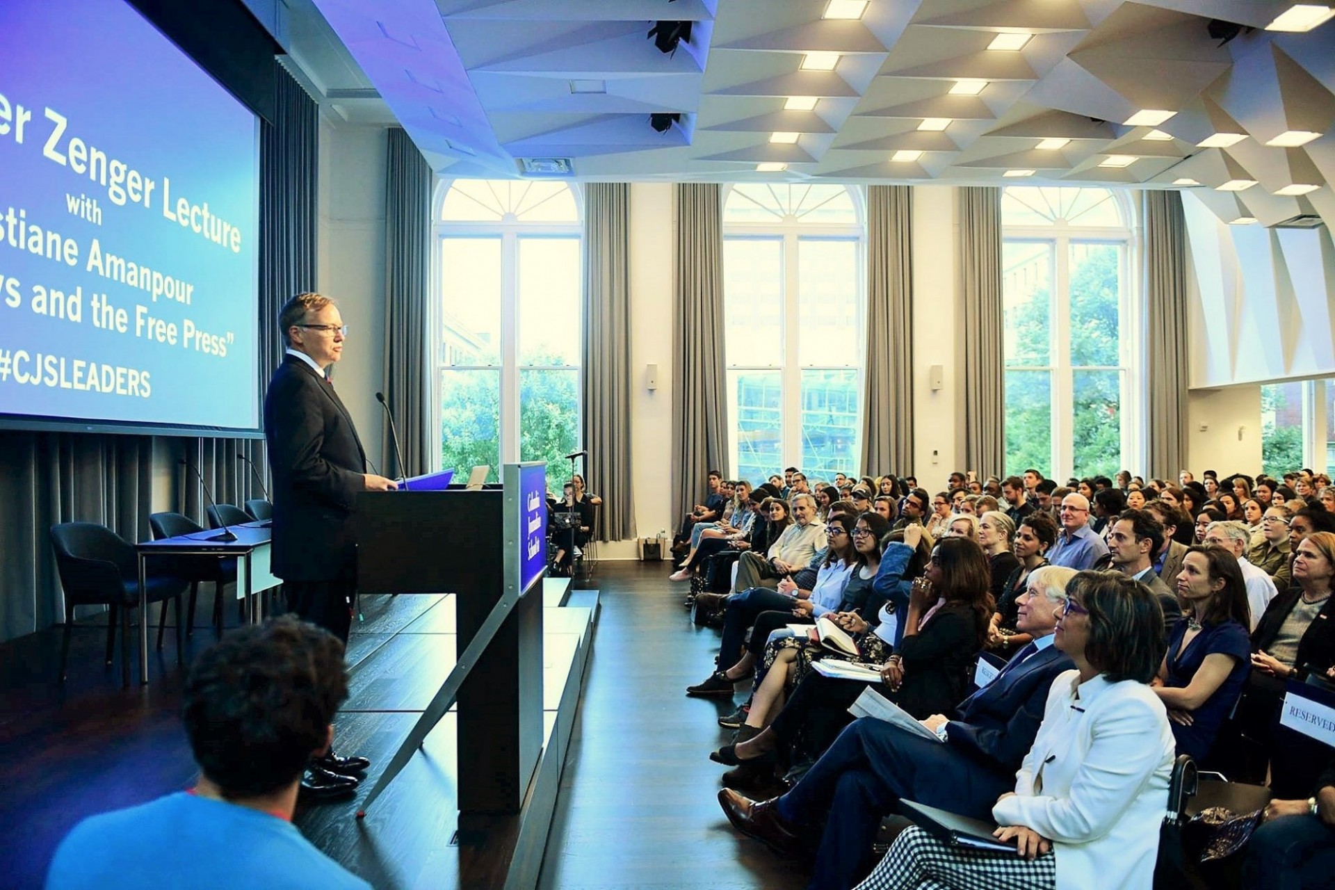 Man in grey suit with glasses stands at podium facing an audience. Behind him is a projector screen. The room is lit with blue lights.