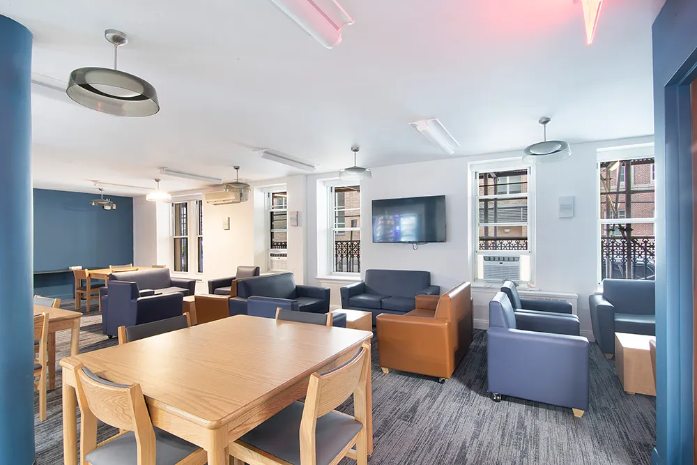 An empty residential lounge with blue couches and lounge chairs and square, wooden study tables.