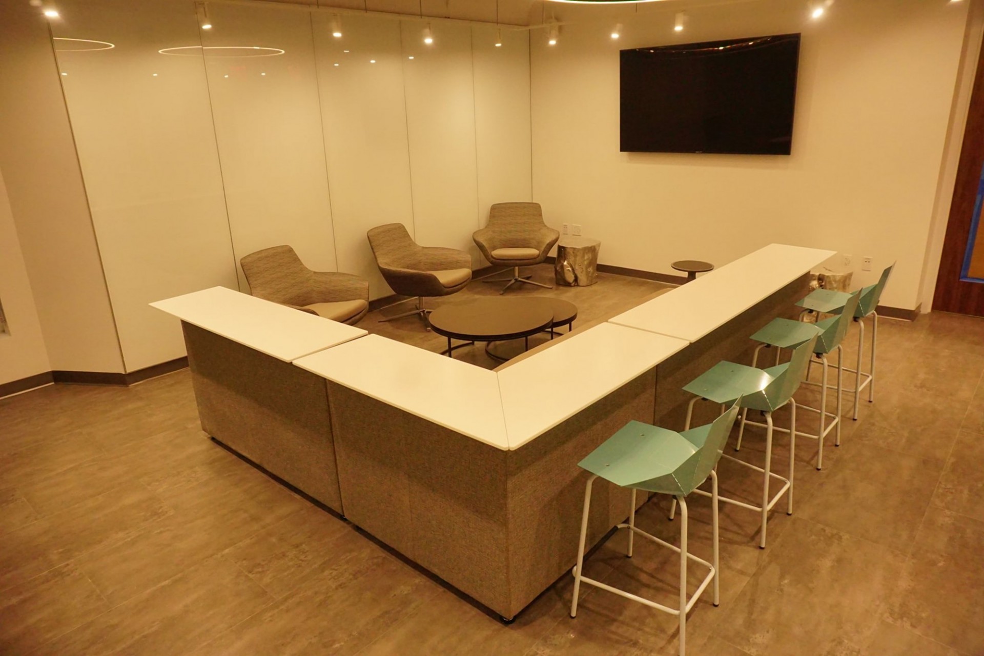 White, high-top counter with 4 light blue stools in a white-walled room with a TV screen on the right wall.
