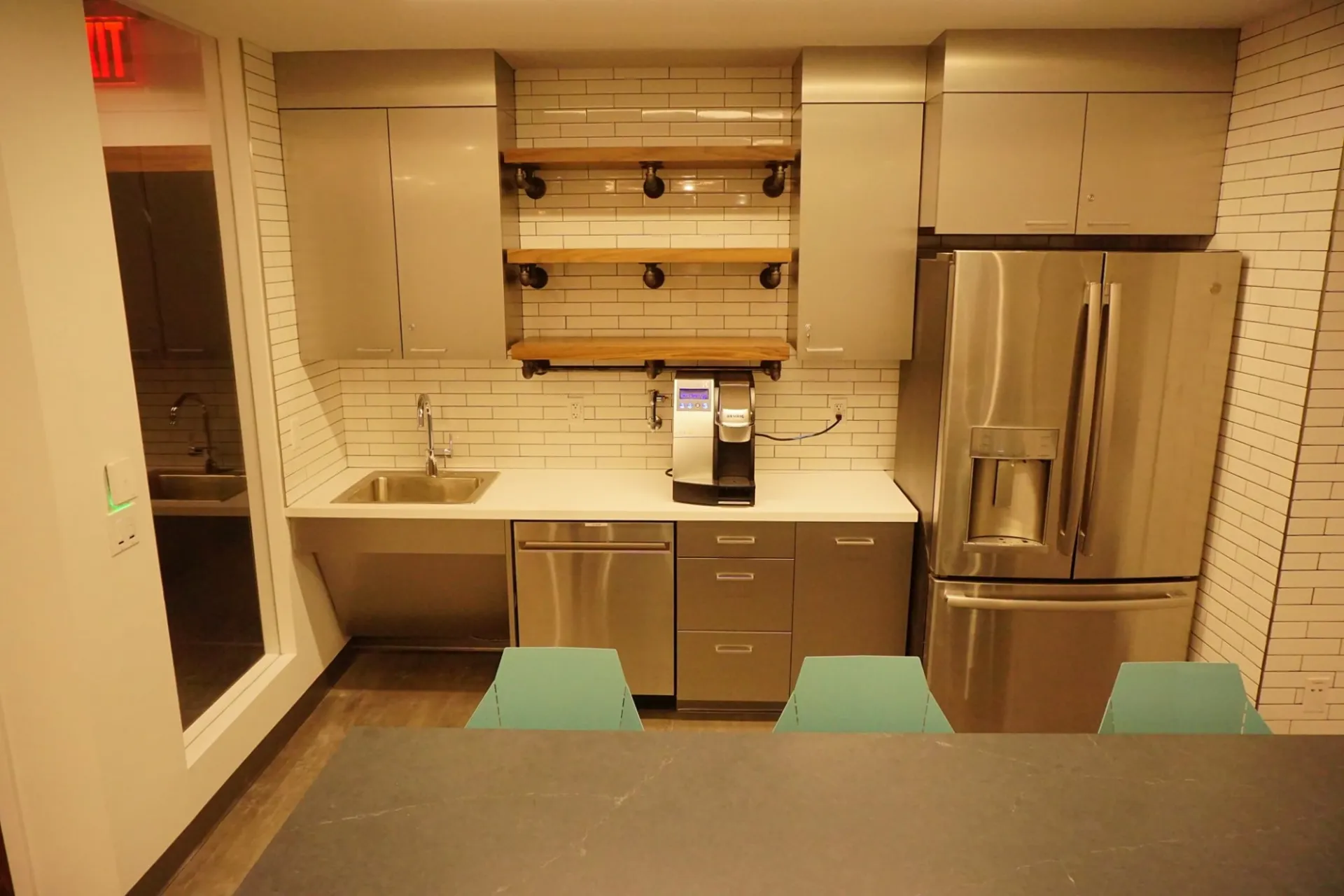 A white, subway tiled kitchen with stainless steel appliances, grey cabinets, and open shelves. At the forefront of the picture is a counter with blue stools.