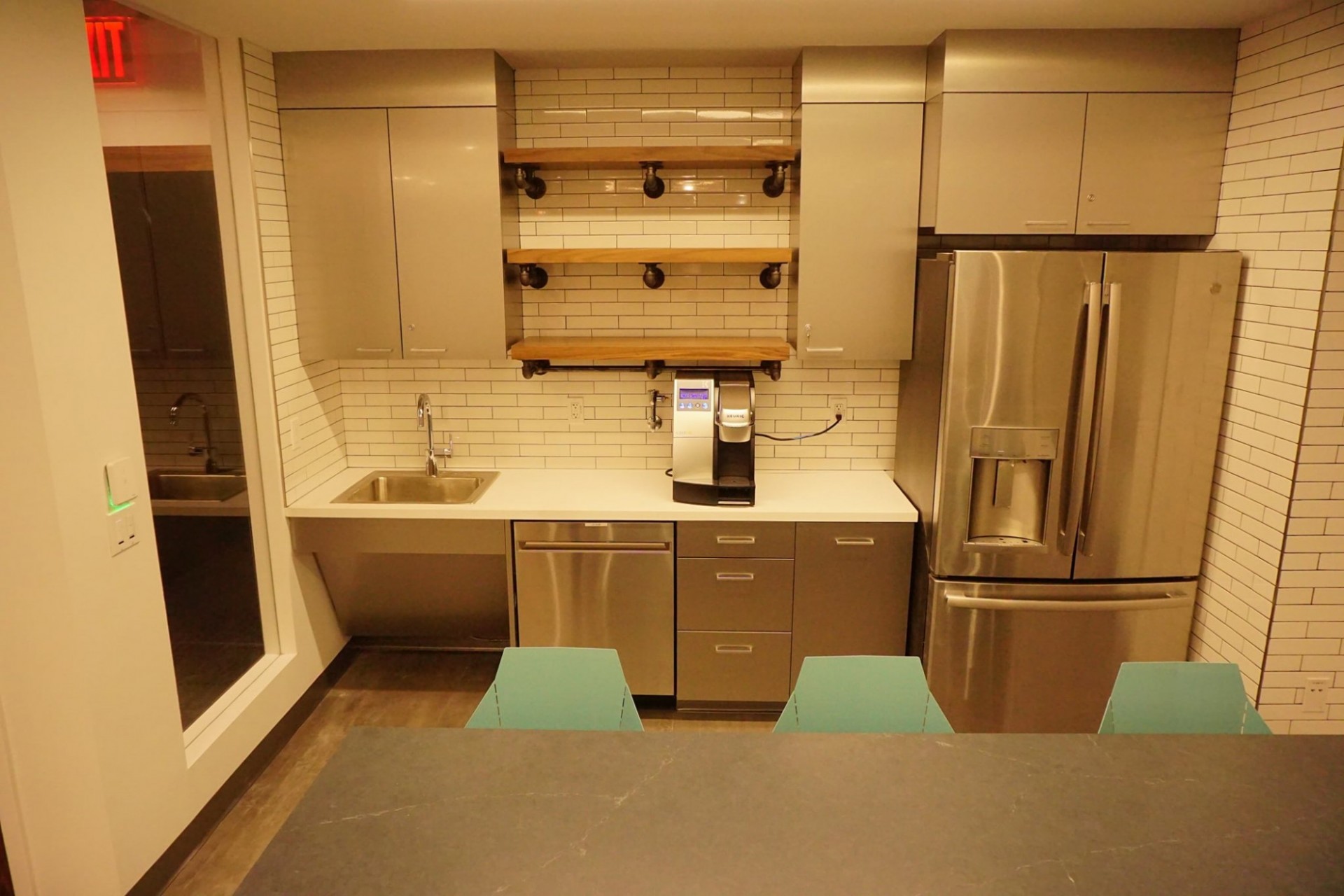 A white, subway tiled kitchen with stainless steel appliances, grey cabinets, and open shelves. At the forefront of the picture is a counter with blue stools.
